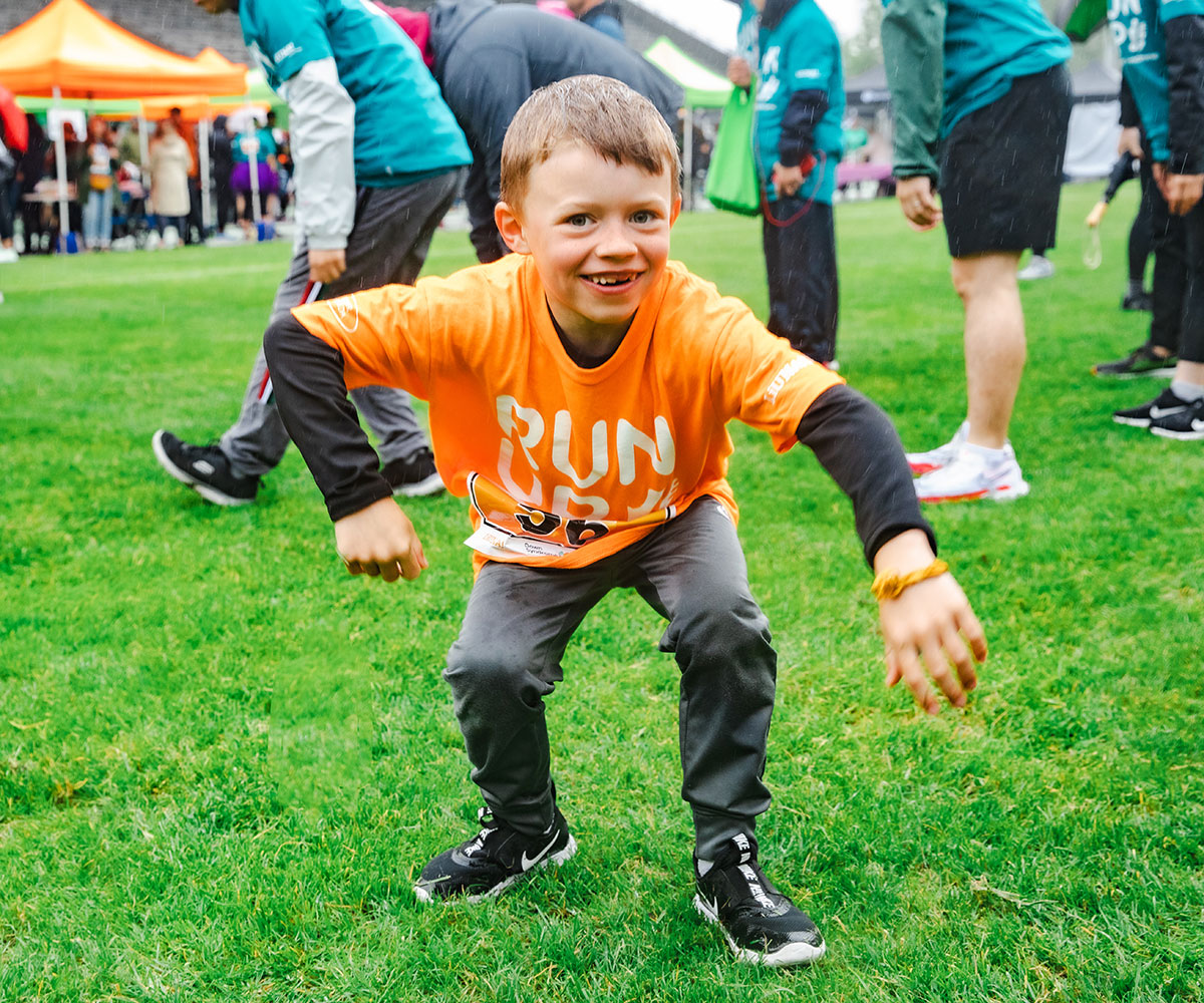 young white boy with brown hair and Down syndrome, wearing orange Run Up for Down Syndrome t-shirt, crouches down while doing warmup exercises