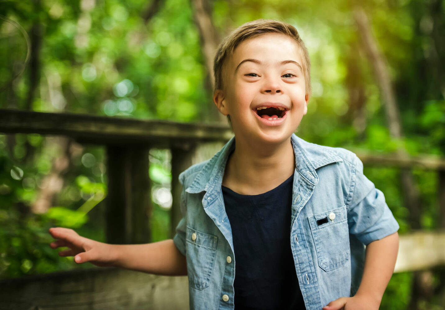 white, blonde haired boy with Down syndrome with black t-shirt and light blue shirt over top smiles on a bridge in the woods
