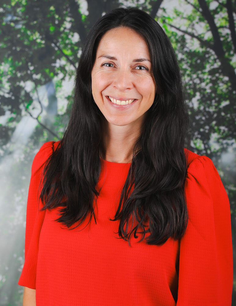 headshot of white woman with long black hair wearing red top against a photo backdrop off sunlight peeking through trees