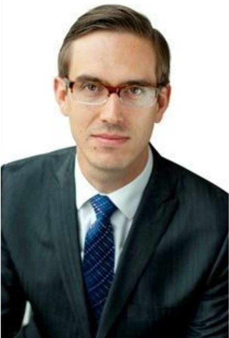 headshot of white man with brown hair, glasses, and grey suit with blue tie