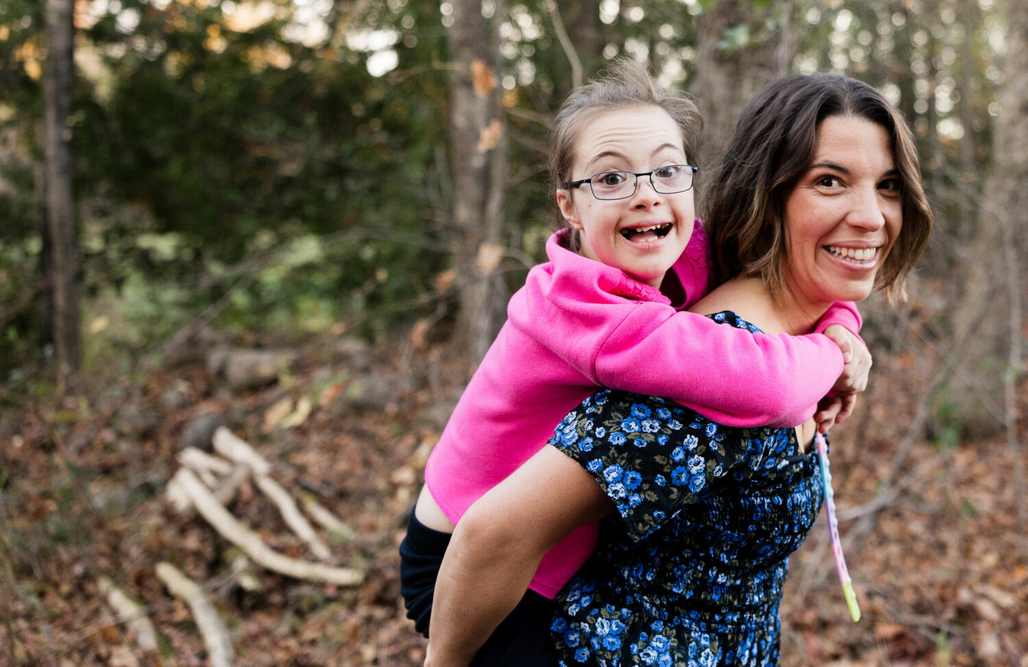 brown haired white woman wearing a blue shirt piggybacks her daughter, a white girl with Down syndrome with brown hair and glasses wearing a pink sweater, in the woods