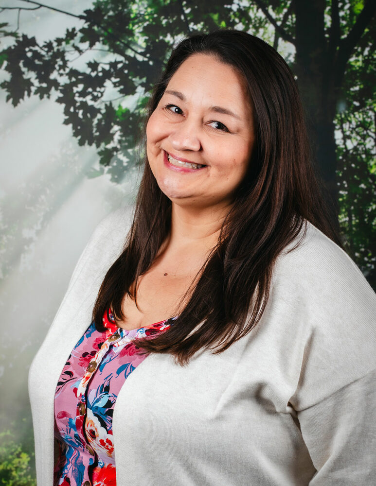 white woman with long brown hair, wearing colourful flowered shirt and light grey sweater, standing in front of a forest picture