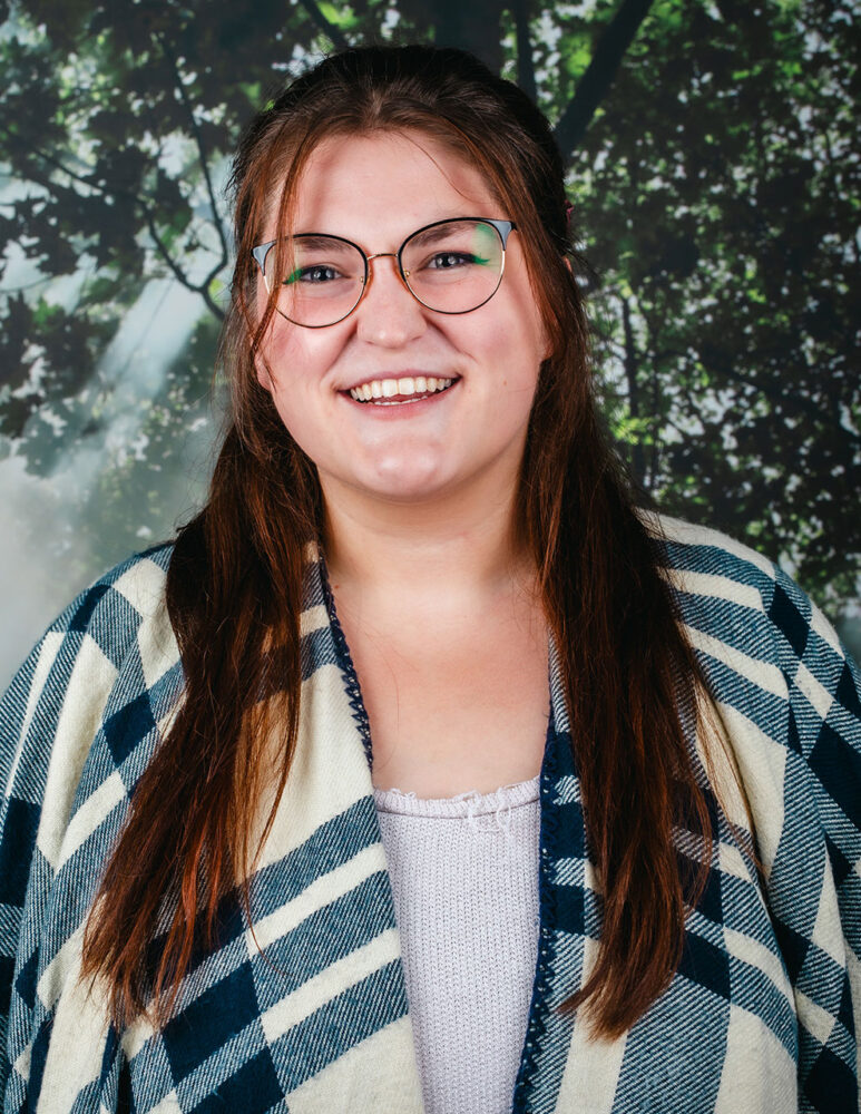 white woman with long brown hair and glasses, wearing green and white patterned sweater over white shirt, standing in front of a picture of a tree