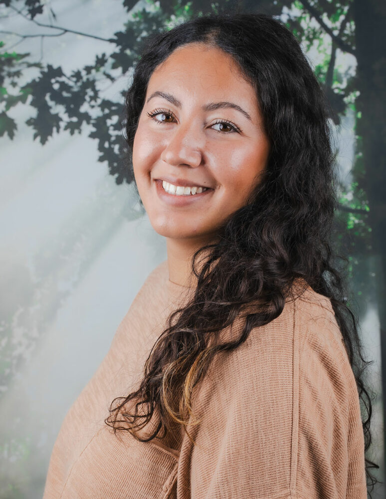 headshot of woman with long, curly brown hair wearing tan top, standing in front of a picture of a tree