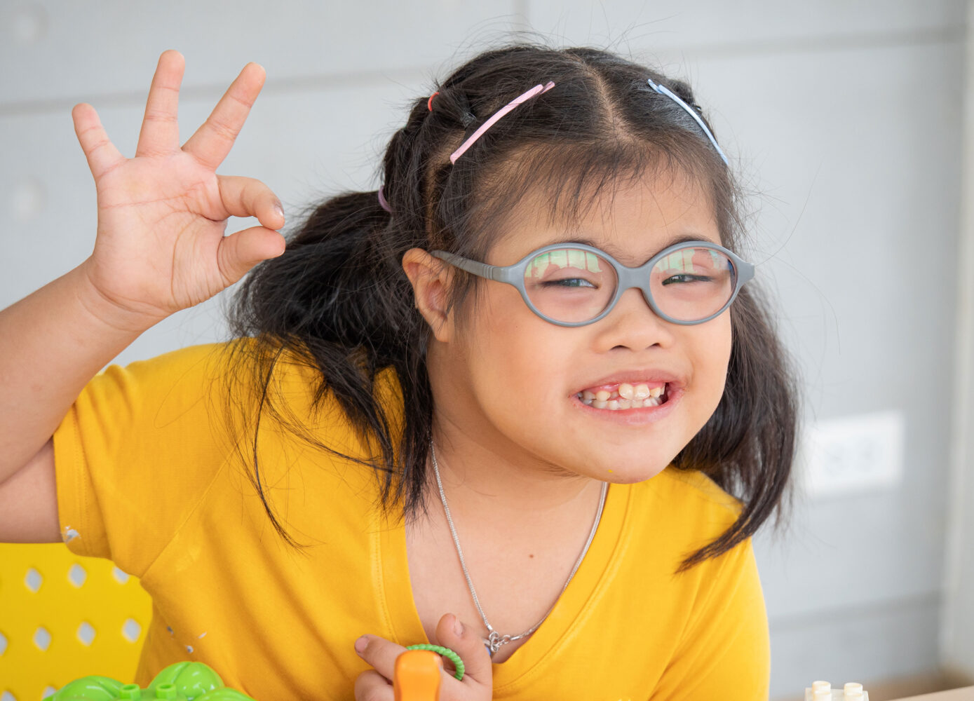 Asian girl with Down syndrome wearing glasses and yellow shirt gives the sign for "okay"