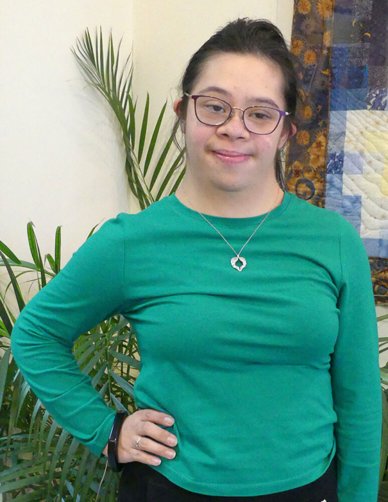 white woman with Down syndrome with dark hair in a ponytail, glasses, long sleeve green shirt, and silver necklace poses in front of a plant