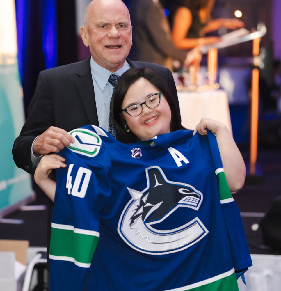 Asian woman with Down syndrome, with long black hair and glasses, holds up blue Vancouver Canucks jersey with #40 on the sleeve, assisted by older white man standing behind her, dressed in a dark suit