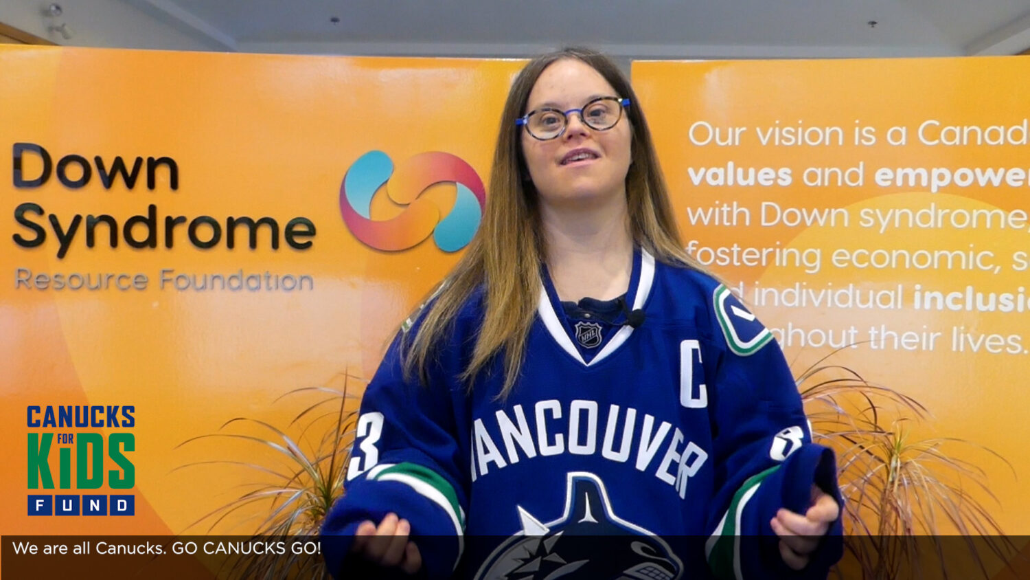 white woman with Down syndrome with long brown hair, wearing glasses and a blue Vancouver Canucks hockey jersey, standing in front of an orange wall with the Down Syndrome Resource Foundation logo