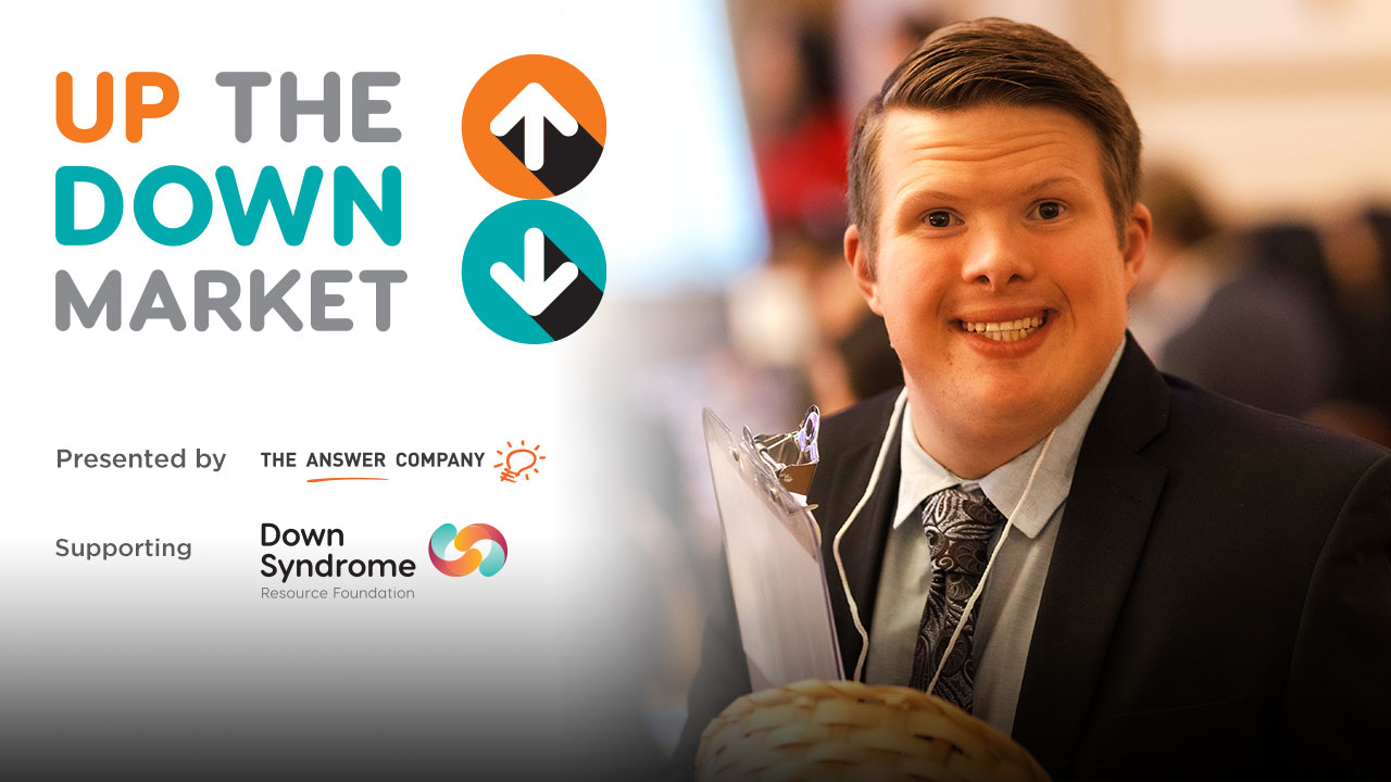 white, brown haired man with Down syndrome wearing a dark suit, holding a basket and a clipboard, with logo for Up the Down Market, presented by The Answer Company and benefiting the Down Syndrome Resource Foundation